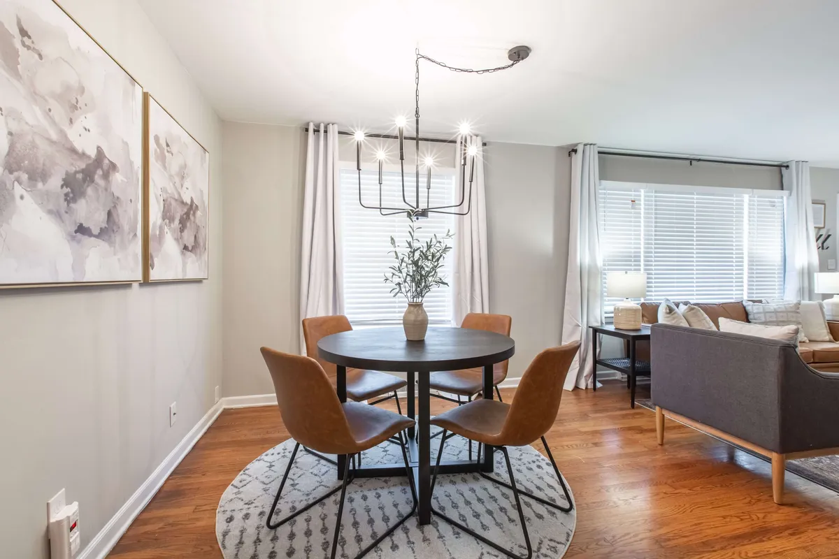 A stylish dining area featuring a central table, contemporary chandelier, wall artwork, and soft window treatments. The space is set up for both intimate dinners and lively gatherings, reflecting versatility for meals or game nights.