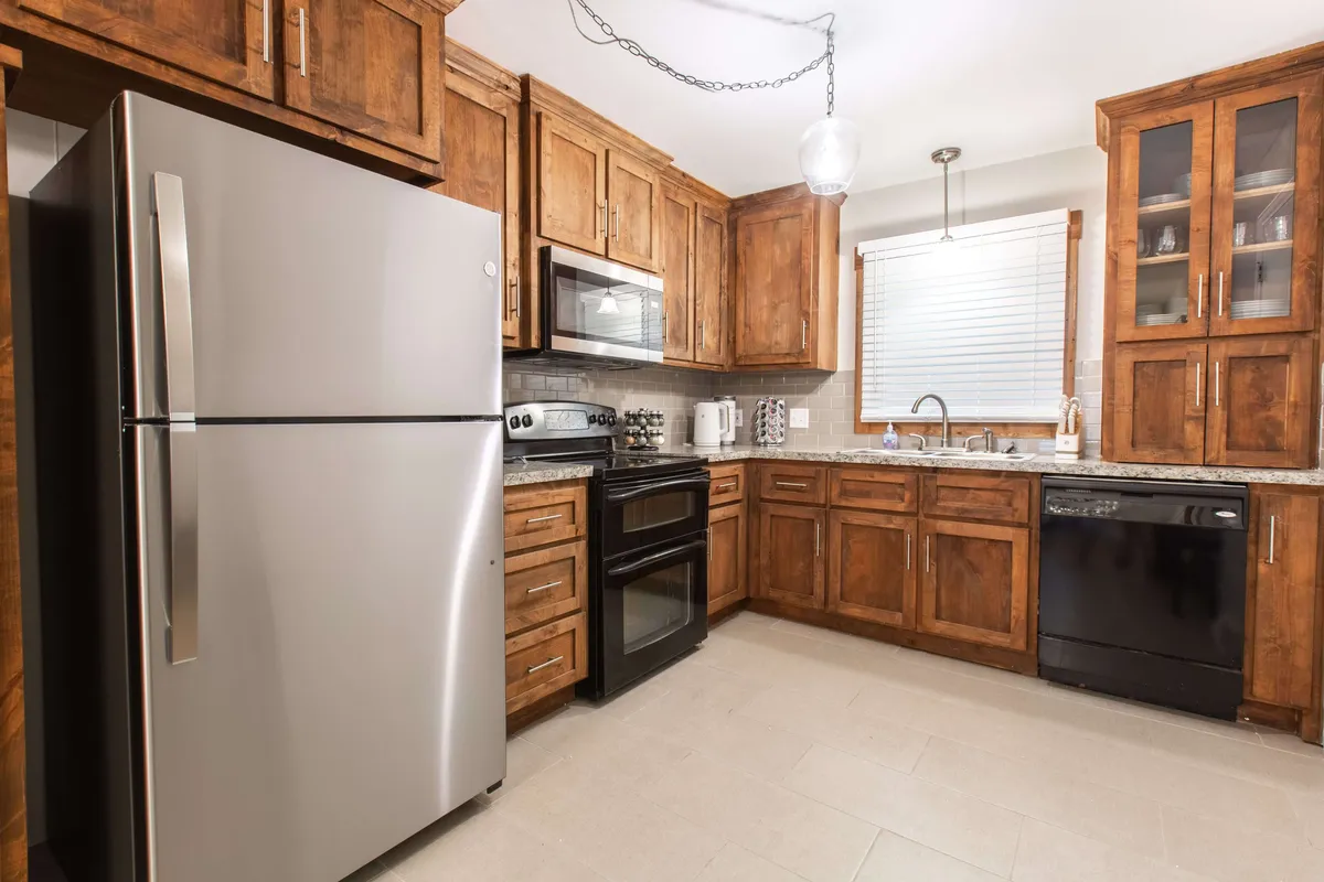 This image presents a cozy, well-equipped kitchen with stainless steel appliances, dark wooden cabinetry, granite countertops, and a window with blinds. The space is inviting, designed for culinary adventures and family gatherings, offering ample room for meal preparation and group interaction.