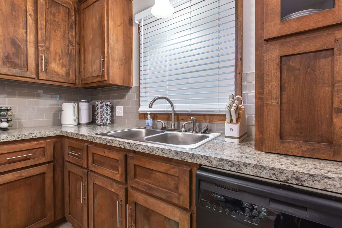 A warm, inviting kitchen with granite countertops, a double sink, and wooden cabinets. Sunlight filters in through window blinds, illuminating a space that feels ready for meal preparation and everyday living.