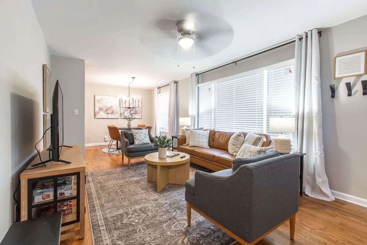 A bright, welcoming living room with a plush sofa, coffee table, television, and large windows letting in natural light. The room feels cozy and is styled for both relaxation and social gatherings. A chandelier and tasteful decor add to the inviting ambiance.