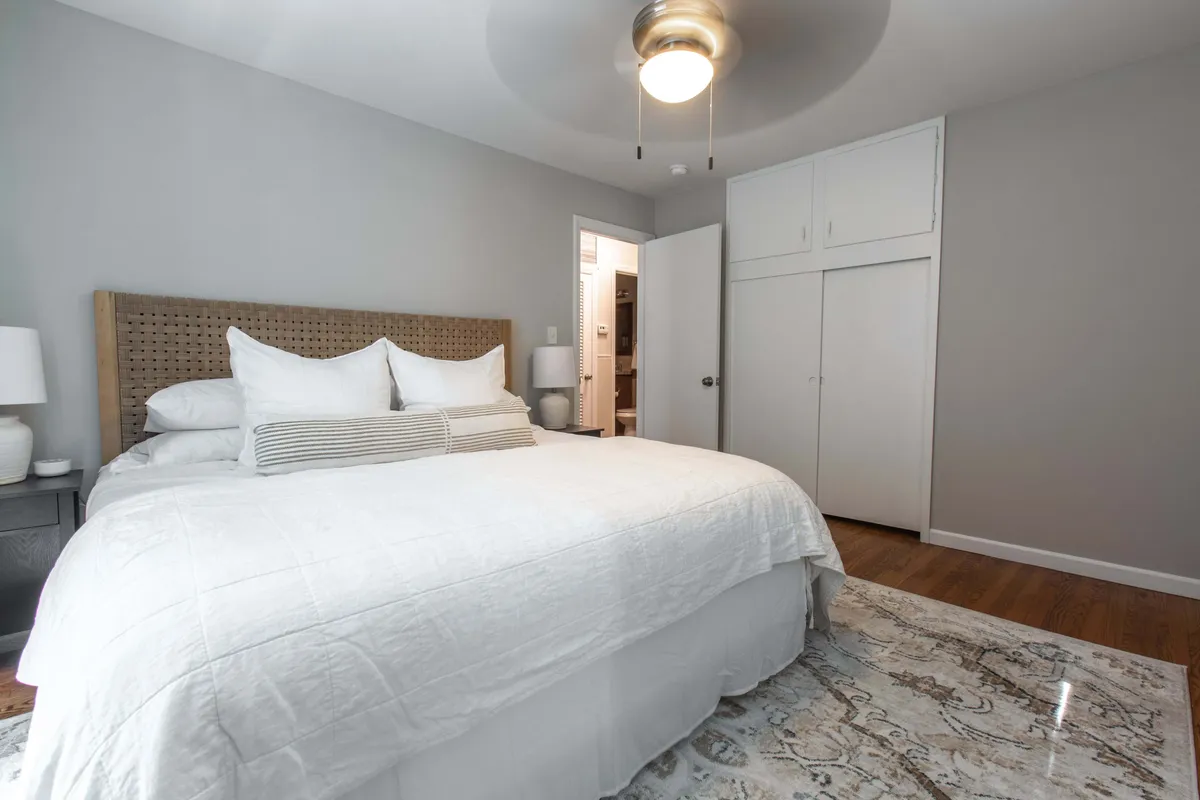 A spacious, comfortable bedroom with a stylish bed adorned with decorative pillows, matching nightstands with lamps, a textured area rug, and an overhead light fixture. The setting suggests an inviting space perfect for reading or unwinding.