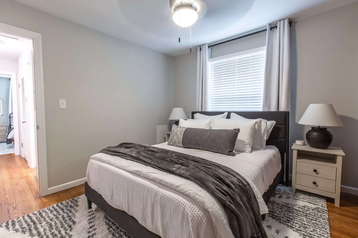 A serene bedroom featuring a neatly made bed with soft linens, a nightstand with a reading lamp, and a sunlit window. The peaceful atmosphere is enhanced by minimal decor, creating a tranquil space ideal for rest and relaxation.